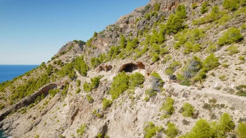 Woman walking along rocky mountain trail near cave and pine trees on Kefalonia coastline