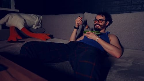 Bearded Guy is Lying on the Couch with Plate of Spaghetti and Chicken and Watching TV Program