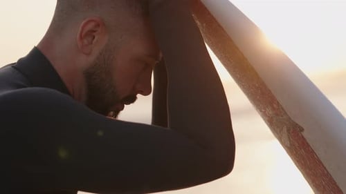 Pensive Surfer Leans on Surfboard at Sunset Contemplating the Ocean