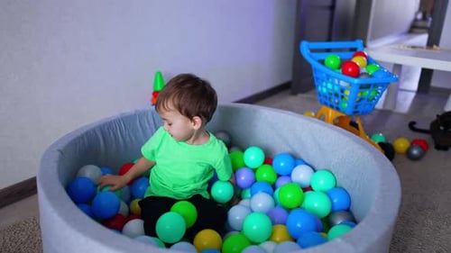 Lovely adorable toddler playing in dry pool at home. Happy kid lies smiling on the colorful balls.