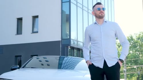 A Handsome Young Business Man is Standing Near His White Car the Business Center in the Background