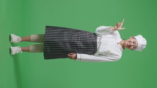 Cuerpo completo de chef asiática sonriendo y apuntando a un lado mientras está de pie en la pantalla verde