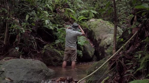 A male traveler hiker stands in a river, creek, stream looking around exploring and discovery with h
