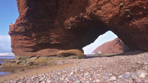 Men Walk on Ocean Coast under Red Rocky Aarch in Legzira Beach in Morocco Summer Travel Backgroun