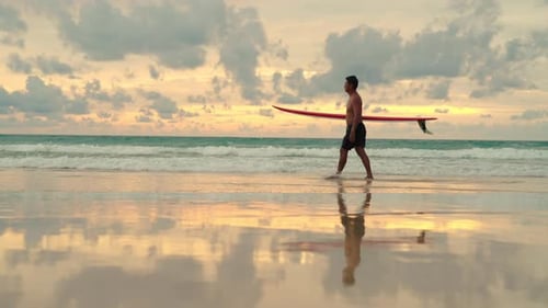4K Asian senior man holding surfboard walking on the beach at summer sunset.